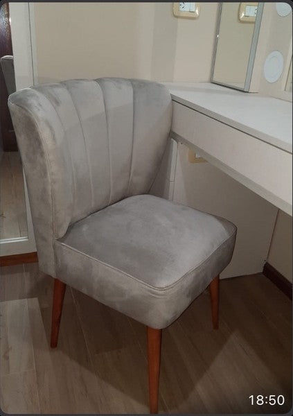 Banquito de tocador gris con respaldo tipo concha y patas de madera clara, de Romy, junto a su tocador
