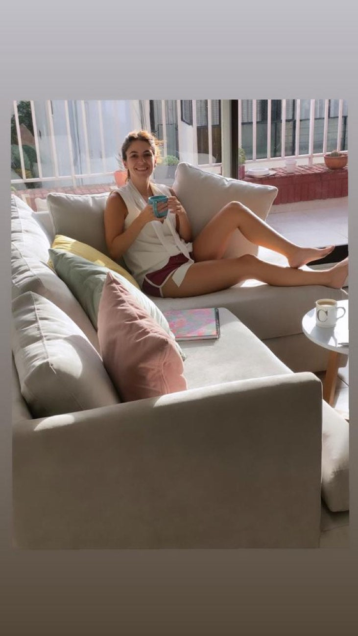 Micaela sentada en su Sillón Esquinero Elton beige, con almohadones pastel, en un living luminoso con balcón.