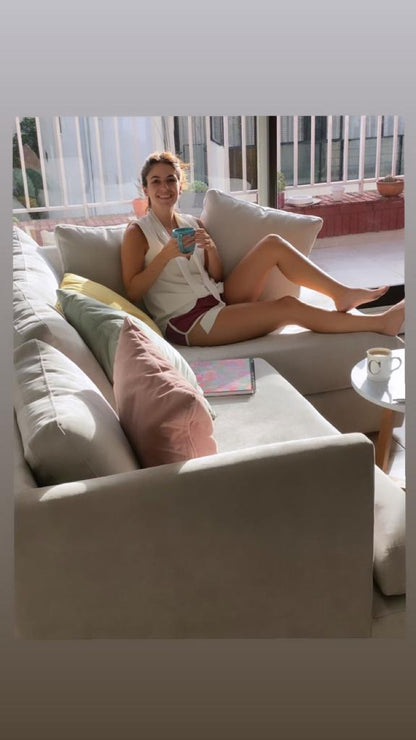 Micaela sentada en su Sillón Esquinero Elton beige, con almohadones pastel, en un living luminoso con balcón.