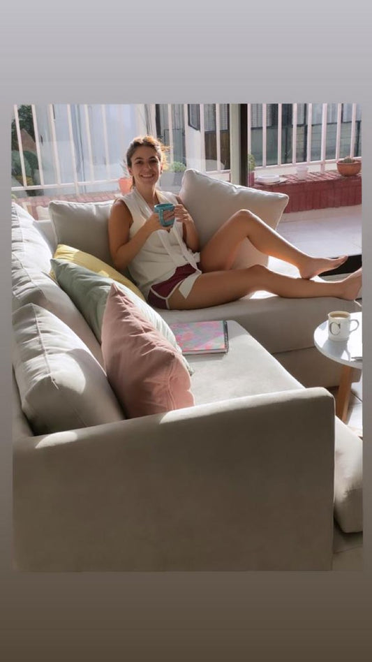 Micaela sentada en su Sillón Esquinero Elton beige, con almohadones pastel, en un living luminoso con balcón.