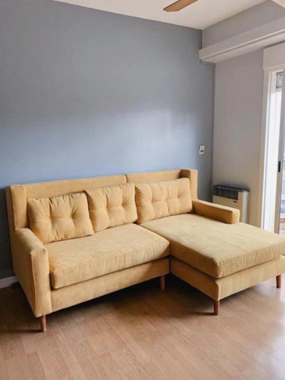 Sillón Esquinero Ragnar tapizado en mostaza, con respaldo capitoné y patas de madera clara, en el living de Melina