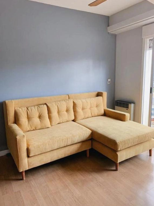 Sillón Esquinero Ragnar tapizado en mostaza, con respaldo capitoné y patas de madera clara, en el living de Melina
