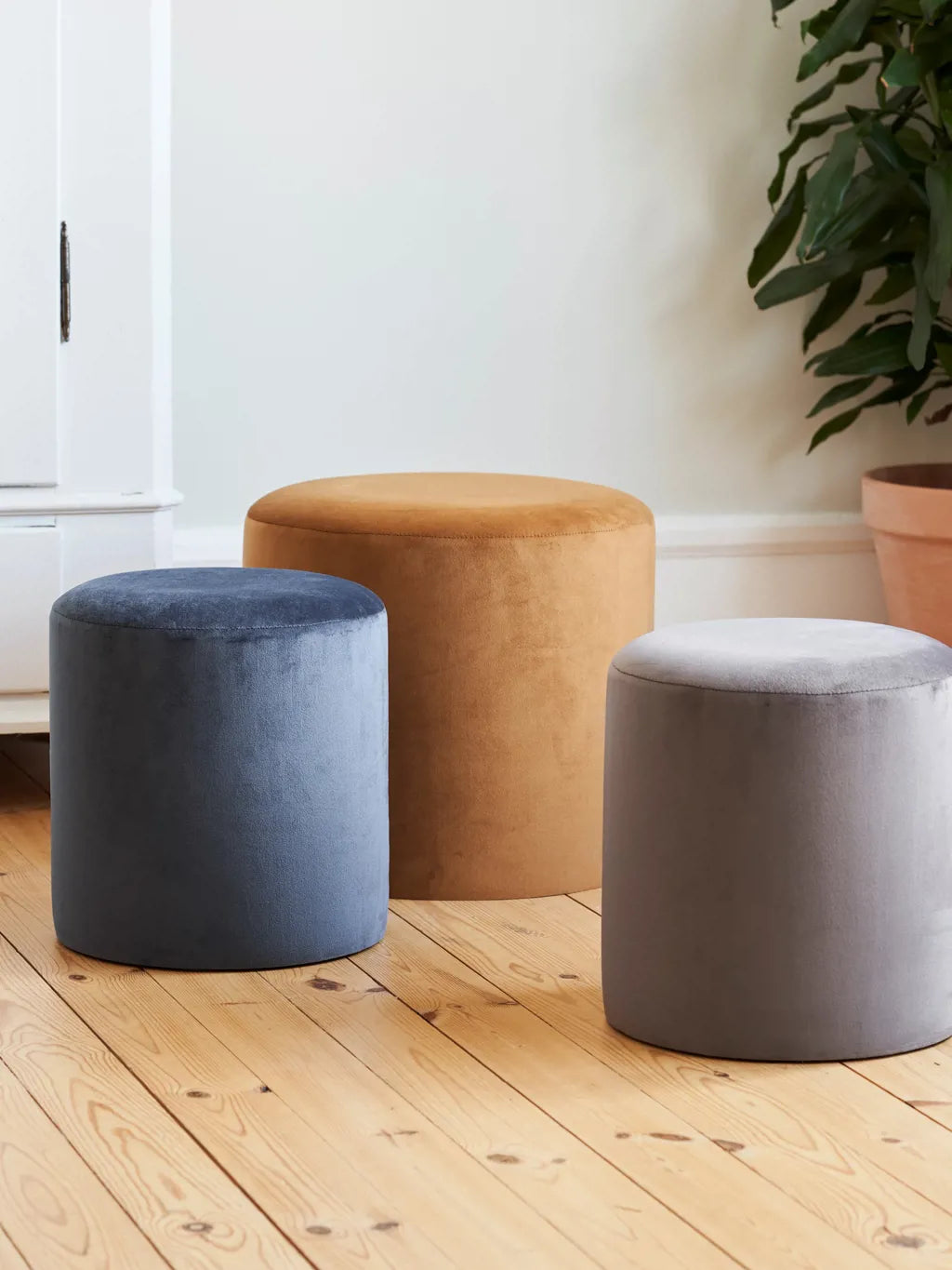 Tres puffs cilíndricos tipo Gemelos en pana azul grisáceo, mostaza y gris, en distintos tamaños, sobre un piso de madera