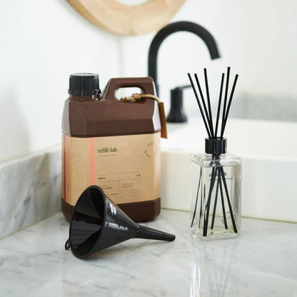 Bidón de difusor Refill Lab con embudo y frasco con varillas negras sobre la mesada de un baño