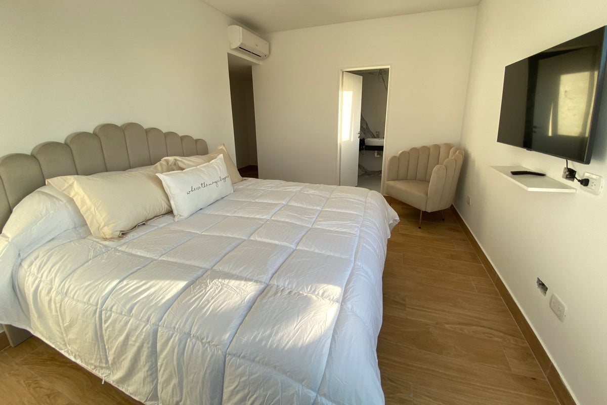 Respaldo Angie beige con paneles redondeados y Sillón Angie beige en forma de concha en un rincón, en el dormitorio luminoso de Flor
