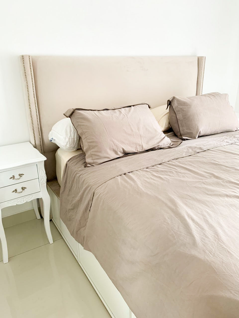Respaldo Emmy beige sin capitoné, con orejas laterales y doble fila de tachas, junto a una mesa de luz bailarina blanca en el dormitorio de Milagros
