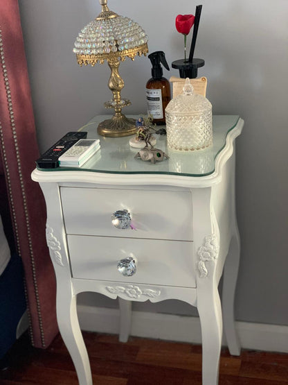 Mesa de luz blanca con tiradores de cristal y lámpara, al lado del Respaldo Emmy rosa