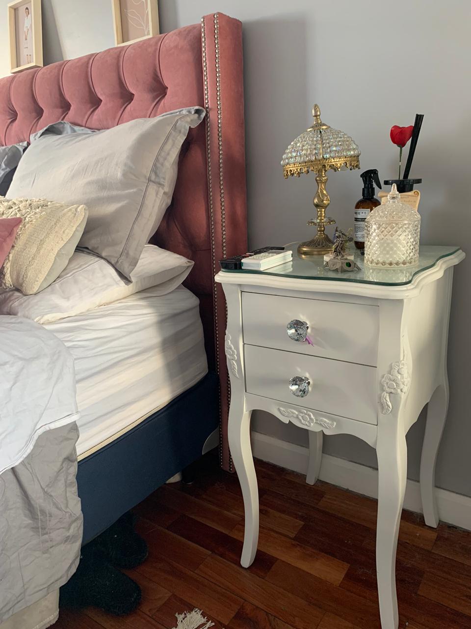 Mesa de luz blanca con lámpara junto al Respaldo Emmy rosa con tachas