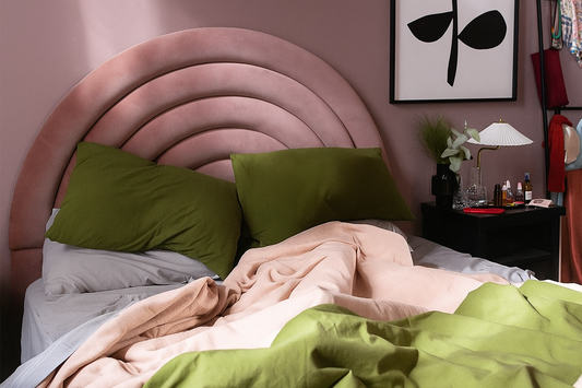 Respaldo de cama tapizado en terciopelo rosa con un diseño semicircular en arcos concéntricos acolchonados. La cama está vestida con sábanas grises y cobijas en tonos rosa pálido y verde oliva, que aportan un contraste suave y natural
