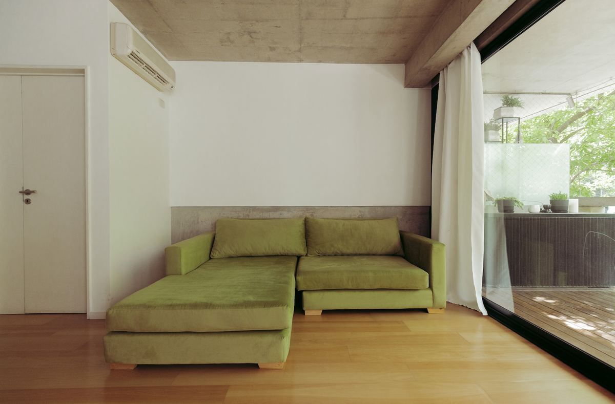 Vista general del Sillón Esquinero Richard verde con chaise longue, en el living de Santiago Colinet con ventanal y piso de madera