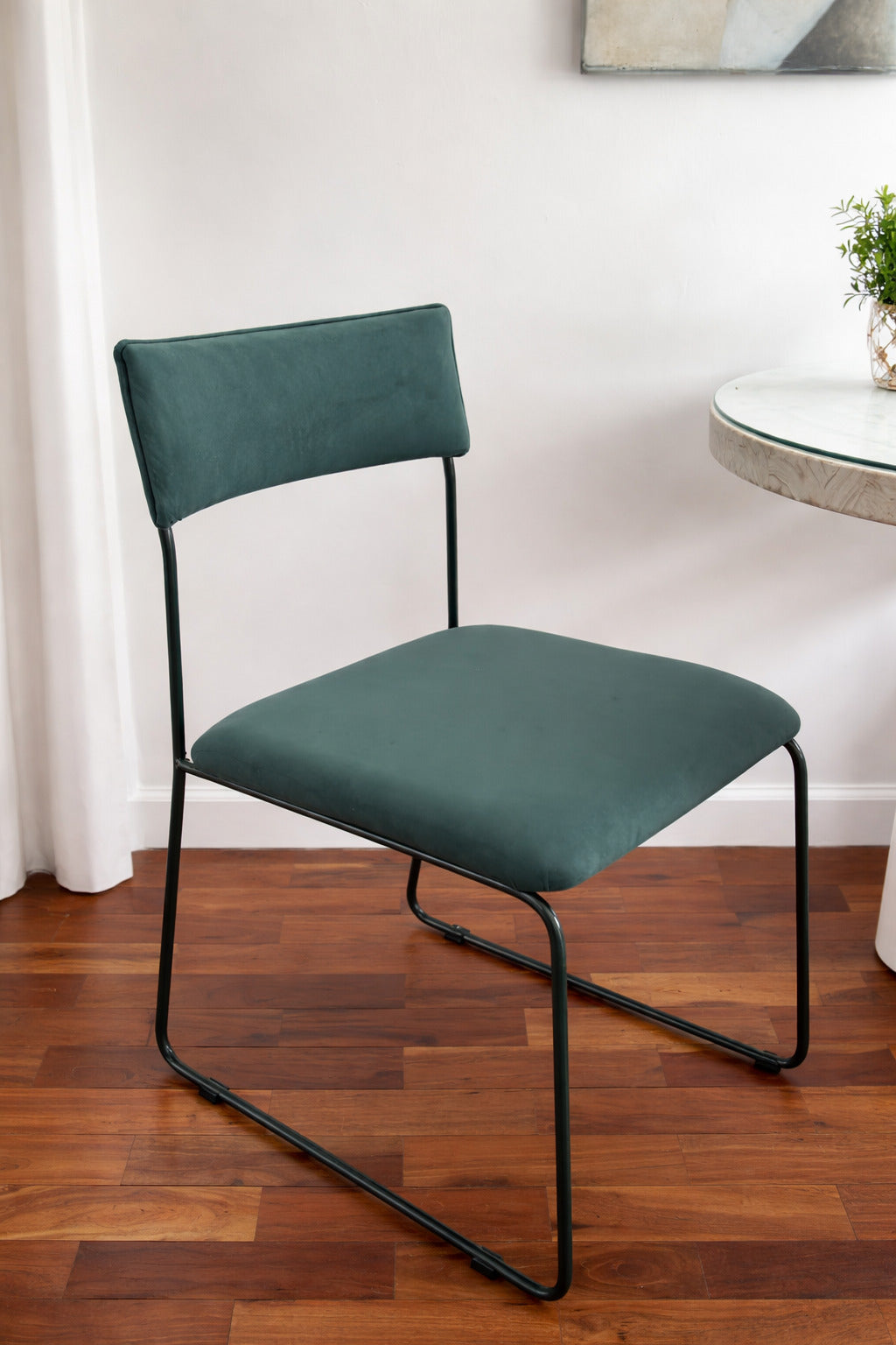 Silla Simple en pana verde azulado con base tipo trineo de caño negro, junto a una mesa de vidrio