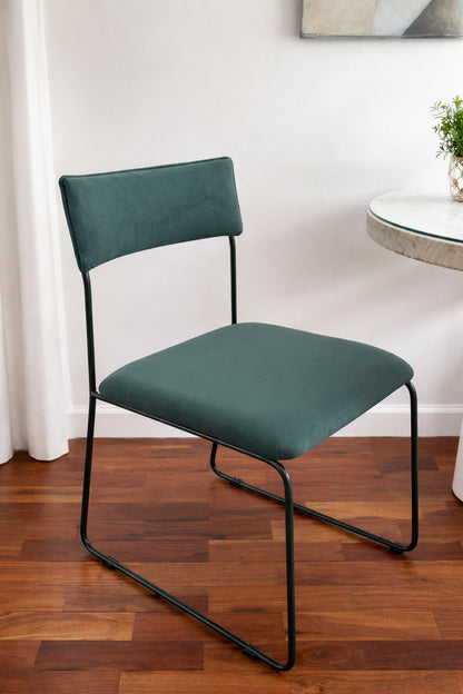 Silla Simple en pana verde azulado con base tipo trineo de caño negro, junto a una mesa de vidrio