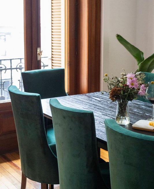 Sillas Amalfi de terciopelo verde alrededor de una mesa de madera oscura con flores, junto a un balcón en San Telmo