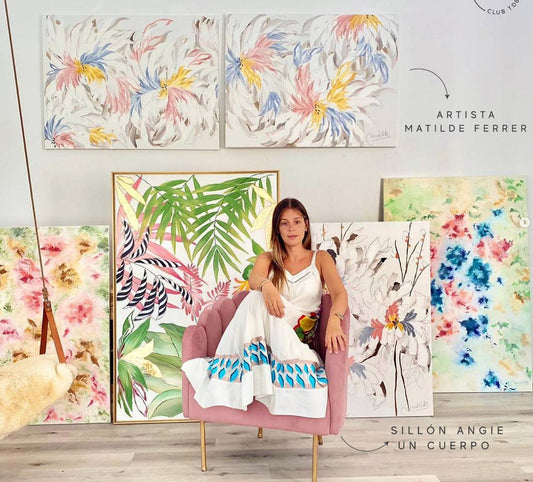 La artista Matilde Ferrer sentada en su Sillón Angie rosa, rodeada de sus cuadros de flores y hojas tropicales