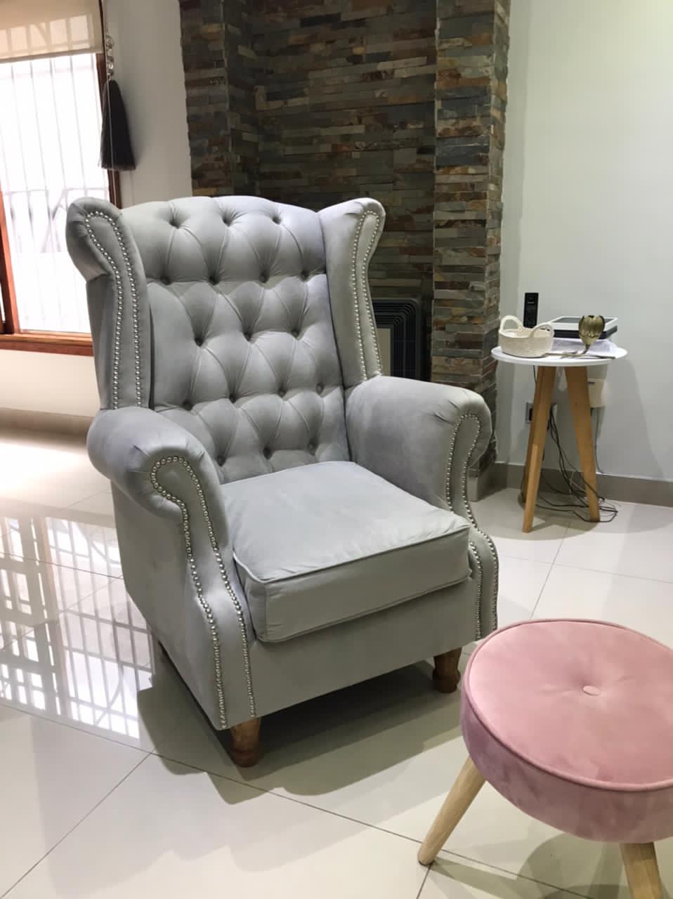 Sillón Elepant gris de respaldo alto capitoné con tachas plateadas, frente a la chimenea de piedra de Sabrina