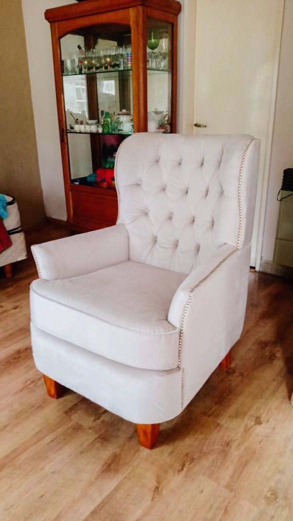 Sillón Esmeralda gris claro con capitoné en el respaldo, tachas plateadas en el contorno y patas de madera, junto a una vitrina en el living de Fabiana