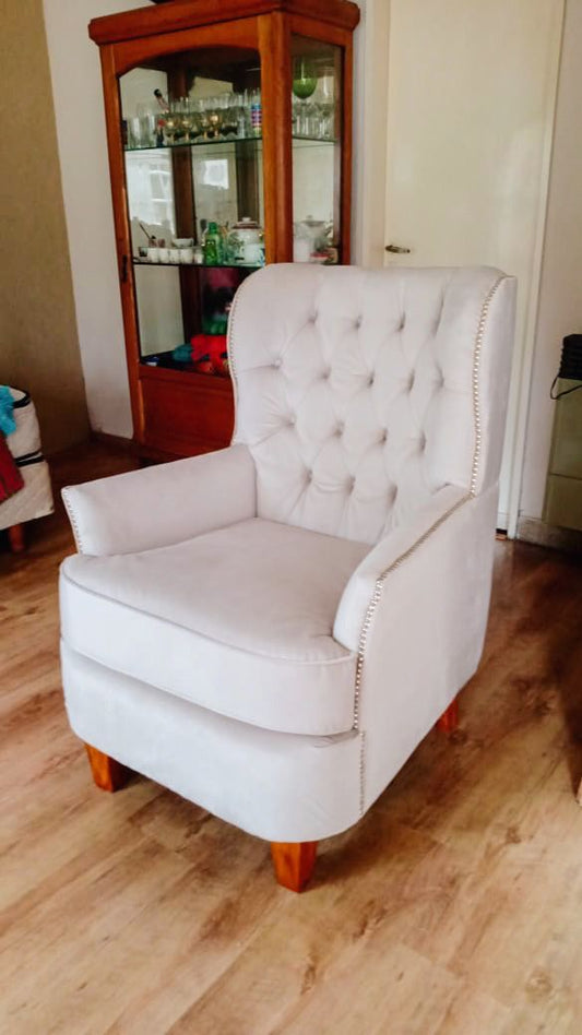 Sillón Esmeralda gris claro con capitoné en el respaldo, tachas plateadas en el contorno y patas de madera, junto a una vitrina en el living de Fabiana
