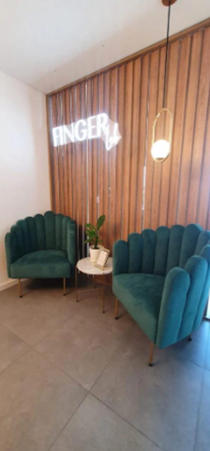 Sillón Angie de un cuerpo y Sofá Angie de dos cuerpos en pana verde inglés con patas doradas, enfrentados junto a una mesa de mármol en la sala de espera de un salón de belleza con pared de listones de madera