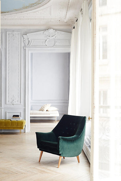 Sillón individual Rivona tapizado en verde, ambientado en living con puertas balcón y piso de parqué espiga, diseño retro con respaldo inclinado y patas de madera.