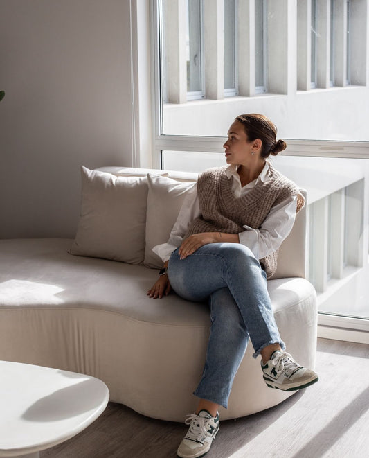 Sillón moderno de forma curva, tapizado en tono claro, con tres cojines decorativos, ubicado junto a una ventana grande que permite la entrada de luz natural, acompañado de plantas en macetas que aportan frescura al espacio