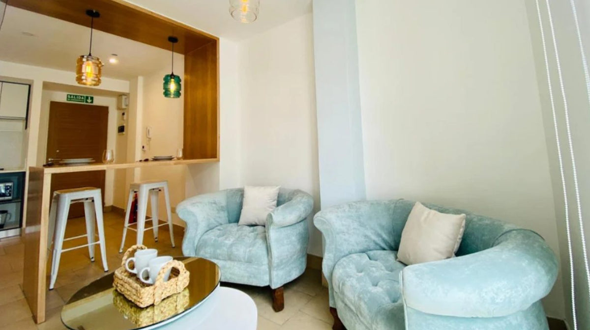 Dos sillones Brigitte tapizados en celeste frente a la barra de la cocina, con mesa baja de vidrio y almohadones blancos