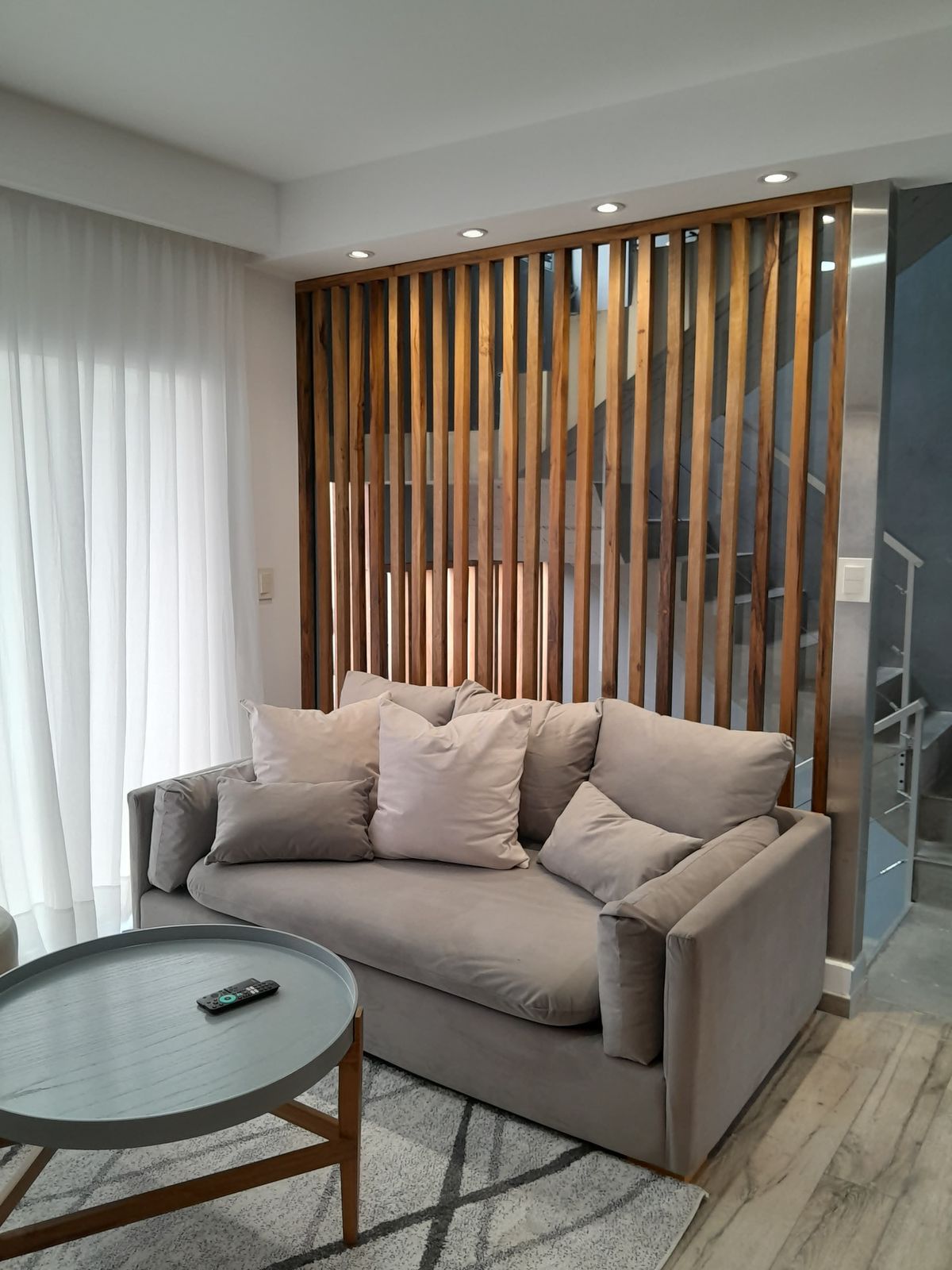 Sofá a medida gris claro con apoyabrazos anchos y almohadones sueltos, frente a un separador de listones de madera, en el living diseñado por Arquitectura SN