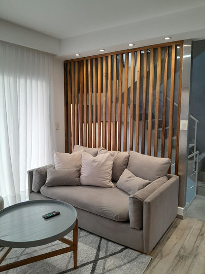 Sofá a medida gris claro con apoyabrazos anchos y almohadones sueltos, frente a un separador de listones de madera, en el living diseñado por Arquitectura SN