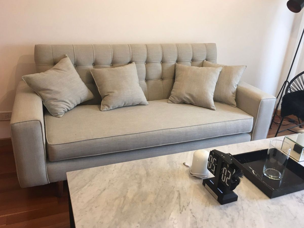 Sofá Alora gris claro con respaldo capitoné de botones, brazos rectos y almohadones al tono, frente a una mesa de mármol en el living de Ludmila