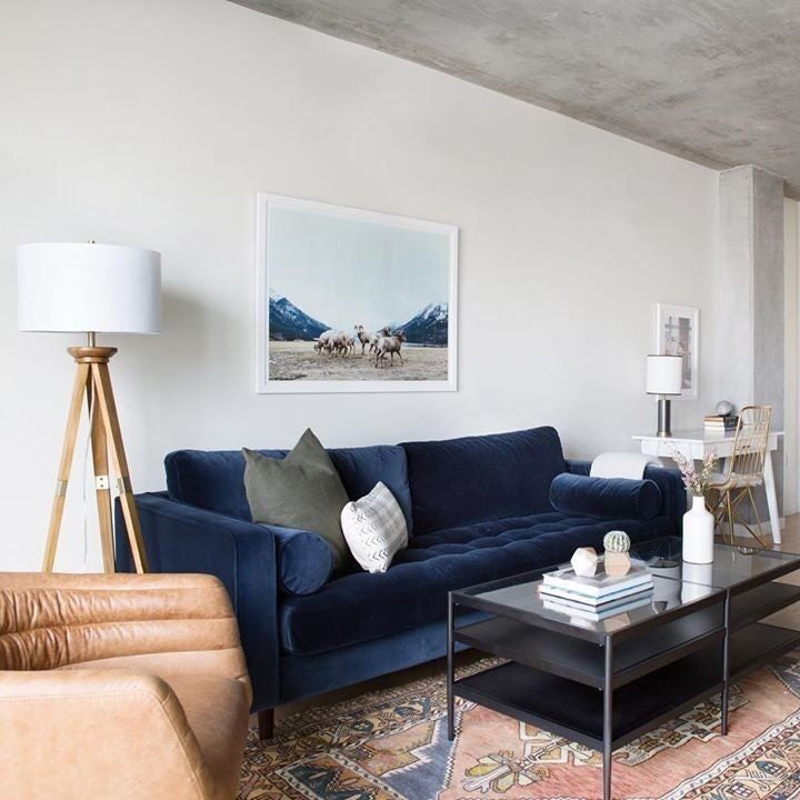 Sofá Bambú en azul marino con botoné en el asiento, junto a un sillón de cuero camel y una alfombra kilim, bajo el techo de hormigón visto del living de Lorena