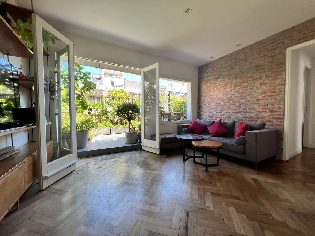 Sofá Bambú gris con asiento botoné y almohadones rojos, junto a un puff al tono y una pared de ladrillo a la vista, en el living de Antonia