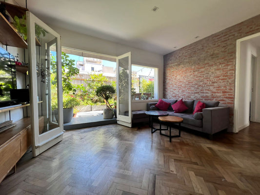 Esquinero Bambú gris en versión sofá más puff, con asiento botoné y almohadones rojos, junto a la pared de ladrillo de Antonia