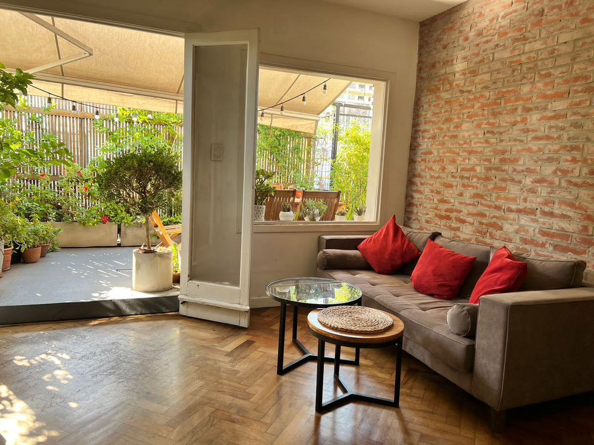 Sofá Bambú gris con asiento botoné y almohadón cilíndrico, frente a la terraza con plantas del living de Antonia