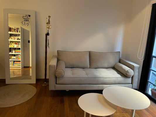 Sofá Bambú gris de dos cuerpos con botoné en el asiento y almohadones cilíndricos, en el living de Micaela junto a un espejo de cuerpo entero