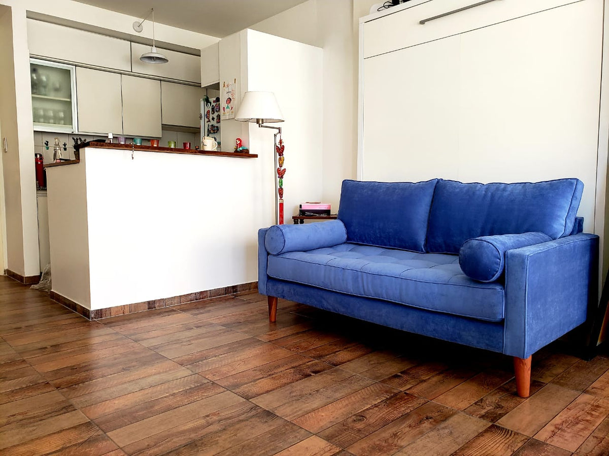 Sofá Bambú en índigo con asiento botoné, almohadones cilíndricos y patas de madera, en el living de Pau