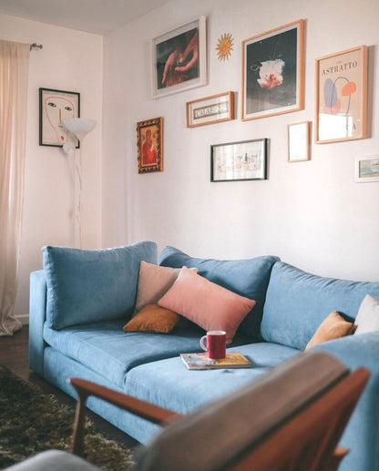 Sofá Elton azul con almohadones rosas y mostaza, bajo una pared de cuadros y láminas