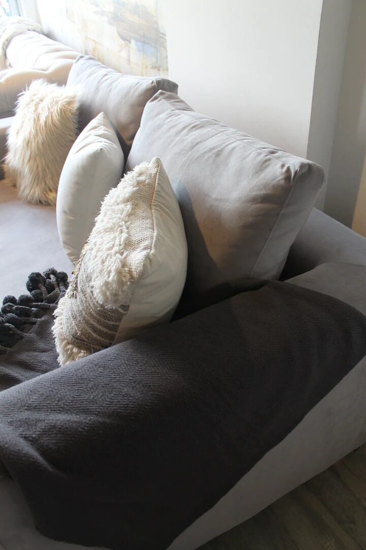Sofá Elton gris con almohadones de lana y pelo y una manta con pompones, en el living boho de Silvana