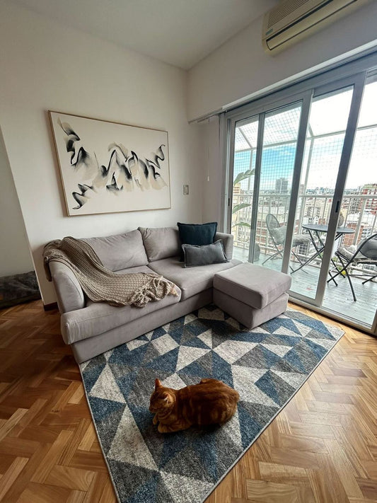 Sofá Elton gris con puff al tono en un living con balcón, alfombra geométrica azul y un gato recostado