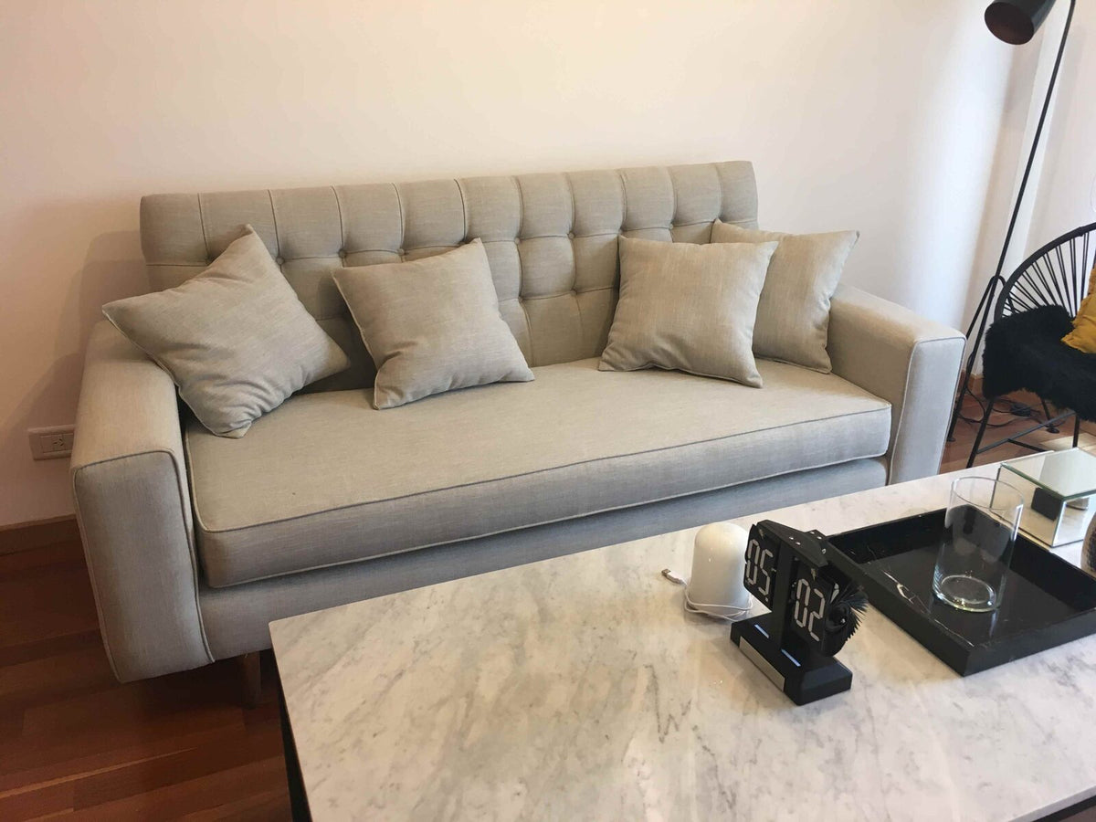 Sofá gris a medida con respaldo capitoné en canales y almohadones al tono, frente a una mesa ratona de mármol blanco en el living de Verónica