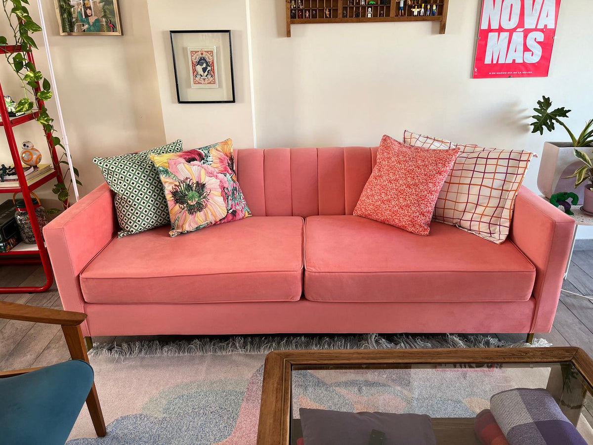 Sofá Mila coral de frente en el living de Mari, con cuatro almohadones estampados y un cuadro con estantería de colección arriba