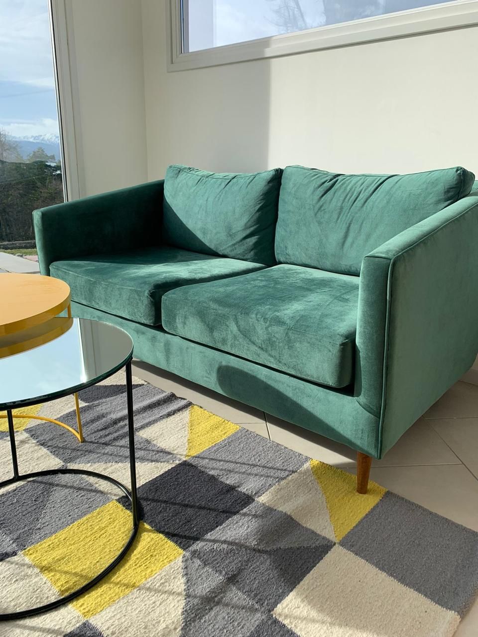 Sofá Philippe de terciopelo verde con apoyabrazos altos y patas de madera, sobre una alfombra geométrica