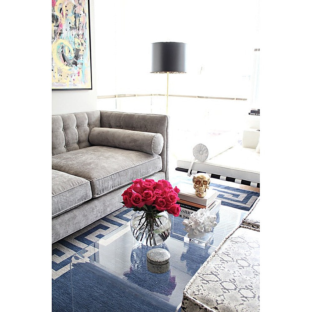 Sofá Valentino gris aterciopelado en vista de tres cuartos, junto a una lámpara de pie y rosas fucsia - Tienda de Banquitos