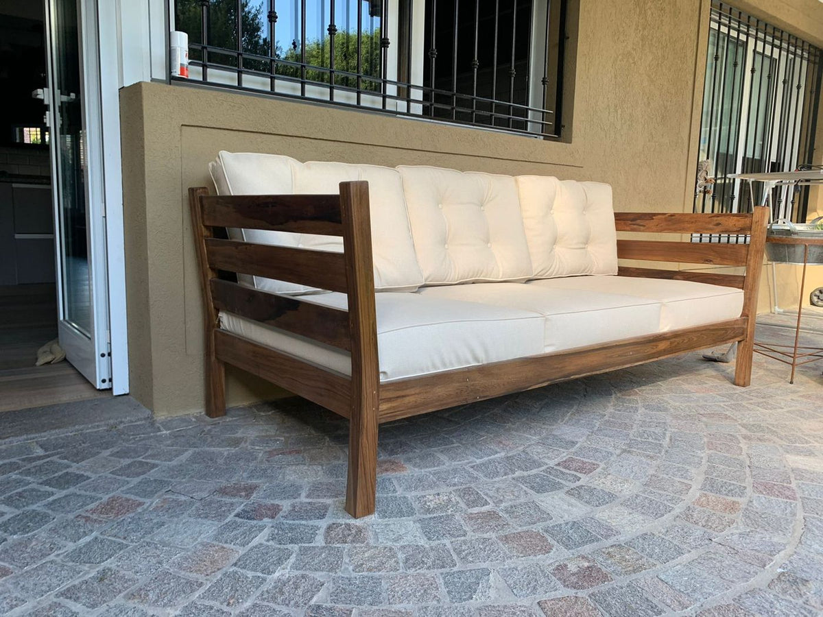 Vista lateral del Sofá Verona de exterior con almohadones crudos, sobre el piso de adoquín de la galería