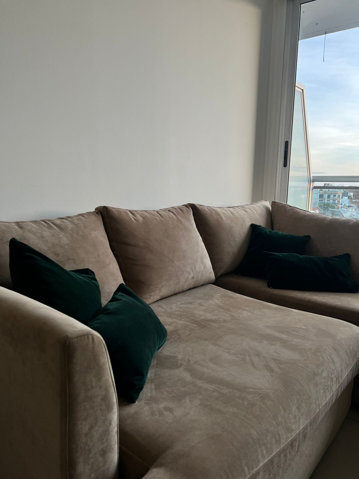 Sofá esquinero en tono beige con cojines a juego, acompañado por un almohadón gris estampado. Frente a él se ubica una mesa de madera con un florero con flores rosadas, en un ambiente luminoso con ventanal.