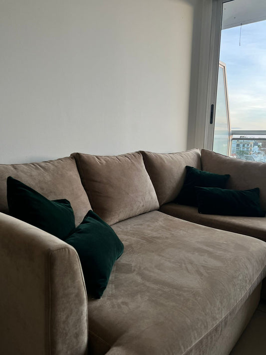 Sofá esquinero en tono beige con cojines a juego, acompañado por un almohadón gris estampado. Frente a él se ubica una mesa de madera con un florero con flores rosadas, en un ambiente luminoso con ventanal.