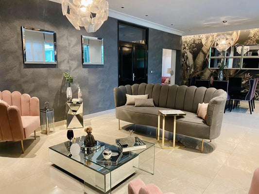 Ambiente elegante con un juego de sillones protagonistas: dos sillones individuales en tono rosa empolvado y un sofá alargado tapizado en gris, todos con un diseño de respaldo acanalado y patas doradas. La disposición de los sillones genera un espacio sofisticado y acogedor, ideal para reuniones o para destacar dentro de la decoración moderna