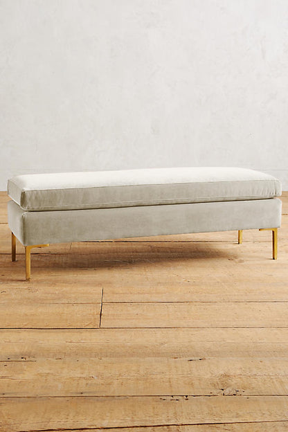 Banqueta de diseño moderno tapizada en crema con patas metálicas doradas. Ideal para pie de cama, recibidores o livings.