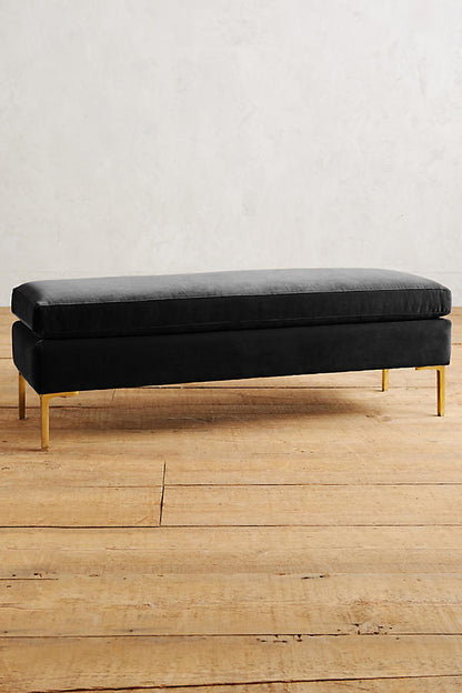 Banqueta de diseño moderno tapizada en negro con patas metálicas doradas. Ideal para pie de cama, recibidores o livings.