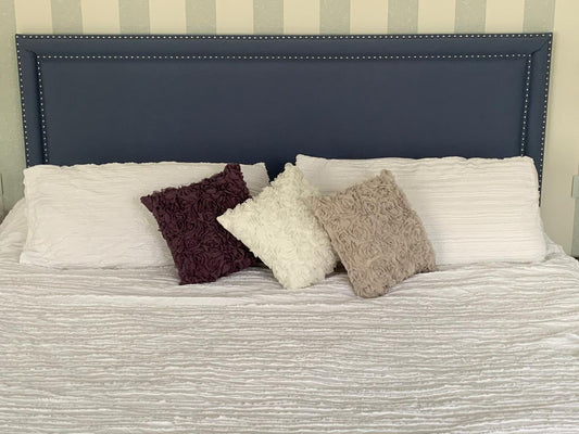 Respaldo de cama rectangular tapizado en color azul marino, con detalle de tachas plateadas en todo el contorno, contrastando con una pared de diseño a rayas verticales.