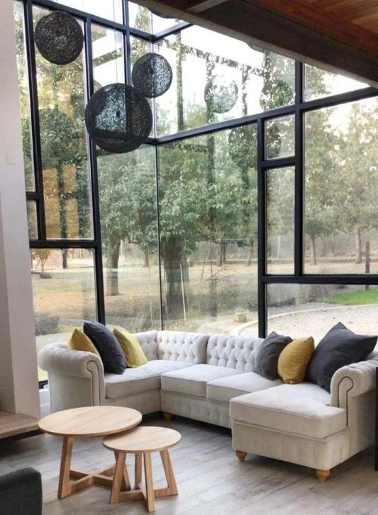 Un sillón esquinero amplio en color beige claro con diseño capitoné, decorado con almohadones grises y amarillos, ubicado en un living con ventanales de vidrio que permiten ver un entorno natural exterior.