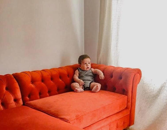 Un amplio sillón en forma de "L" tapizado en terciopelo rojo anaranjado ocupa gran parte de la imagen. El diseño del sillón es clásico, con respaldos y brazos acolchados de capitoné. En una de las esquinas del sillón, se encuentra un bebé pequeño, lo que acentúa la gran escala del mueble. La luz natural entra desde una ventana con cortina blanca al lado derecho, iluminando parte del sillón y del piso de madera.